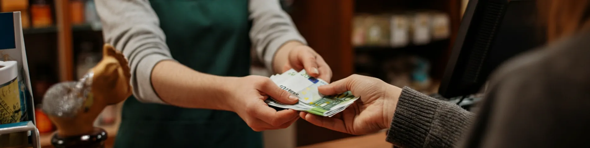 Mano pagando con billetes en euros en una pequeña tienda de barrio, con el tendero detrás del mostrador