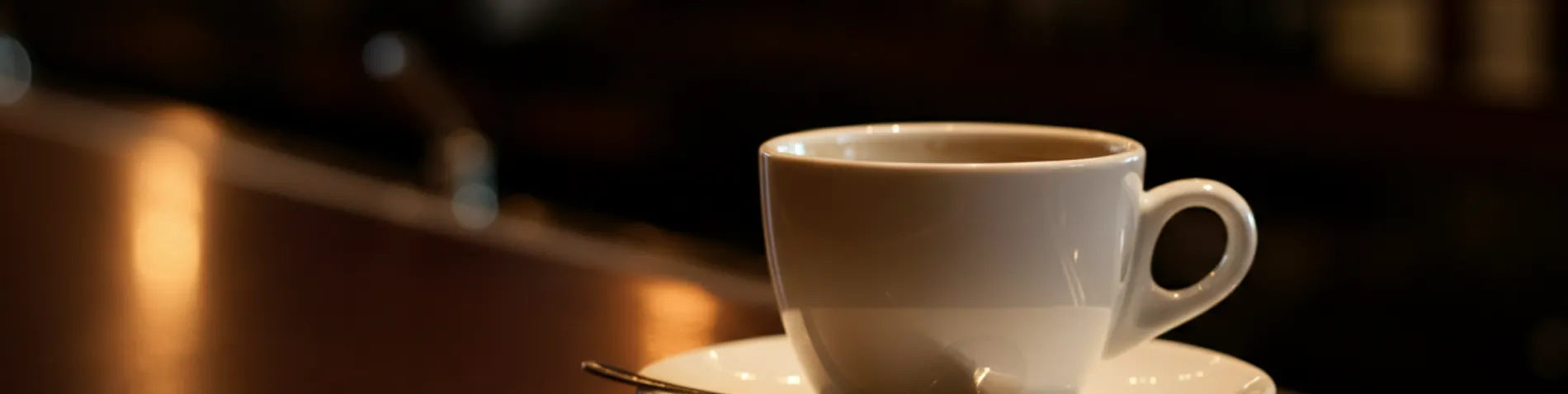 Taza de café en la barra de un bar junto a unas monedas y un platillo, con luz de mañana